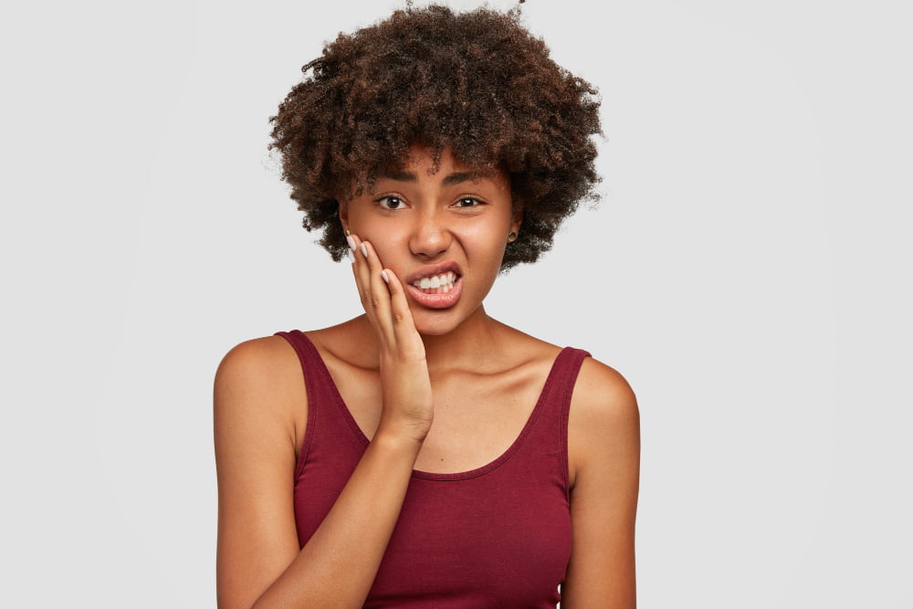 Quanto custa a cirurgia de sisos? 2 displeased beautiful african american woman keeps hand cheeks feels toothache clenches teeth from pain has dark healthy skin crisp hair dressed casually negative facial expressions 2